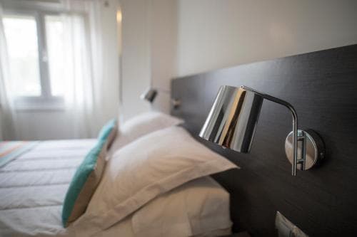 A cozy hotel room features a neatly made bed with soft pillows, a sleek modern wall lamp, and a bright window, creating a serene atmosphere ideal for travelers seeking comfort and relaxation.