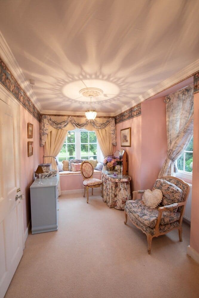A cozy, pastel-pink bedroom with floral furniture, a chandelier casting intricate shadows, and large windows inviting natural light, ideal for a serene getaway or romantic retreat.