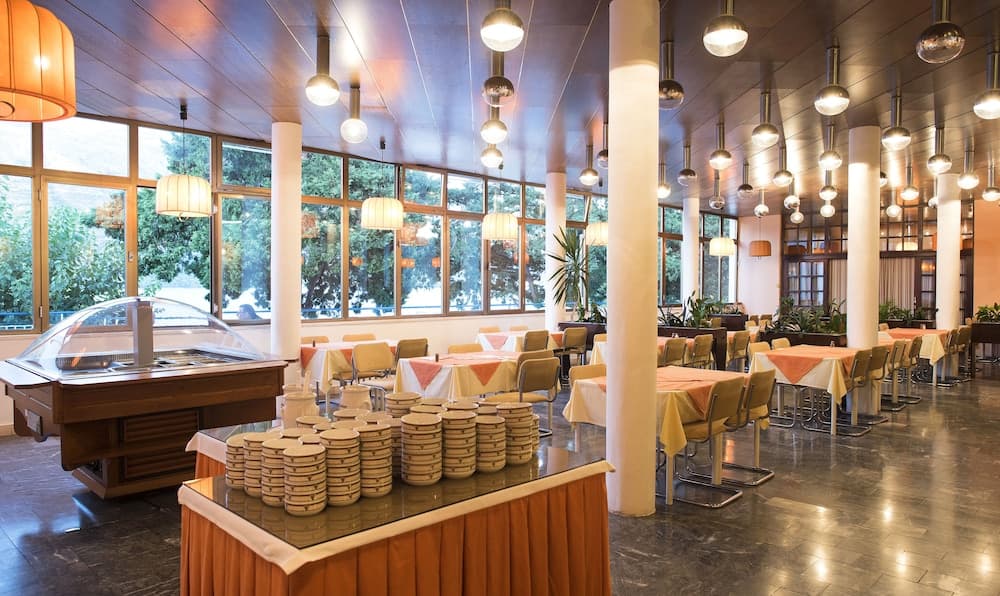 A spacious, well-lit dining hall with large windows overlooking greenery, featuring elegant pendant lights, neatly arranged tables with orange accents, and a buffet setup, ideal for travelers seeking a comfortable and inviting meal environment.