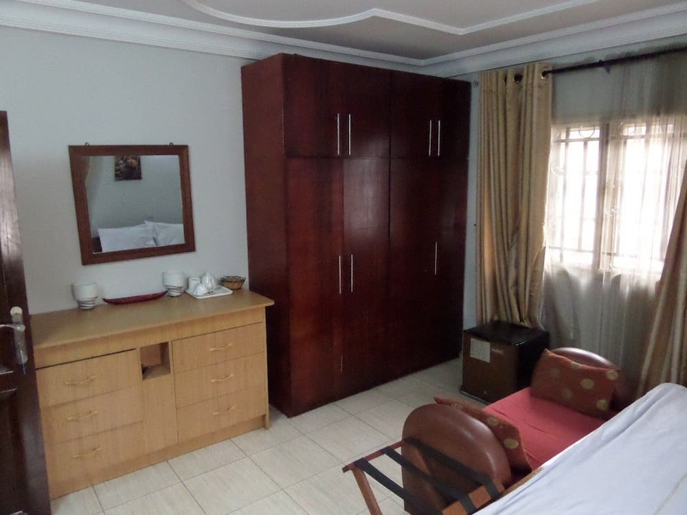 A cozy hotel room features a wooden wardrobe, dresser with tea set, red armchair, and soft lighting through sheer curtains, ideal for travelers seeking comfort and convenience.
