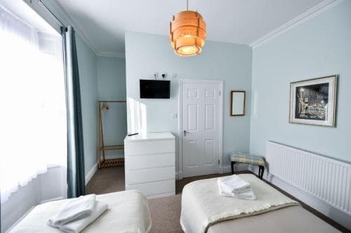 A cozy hotel room with soft blue walls, a neatly made bed, a vintage-style pendant light, and natural light streaming through sheer curtains, ideal for travelers seeking comfort and relaxation.