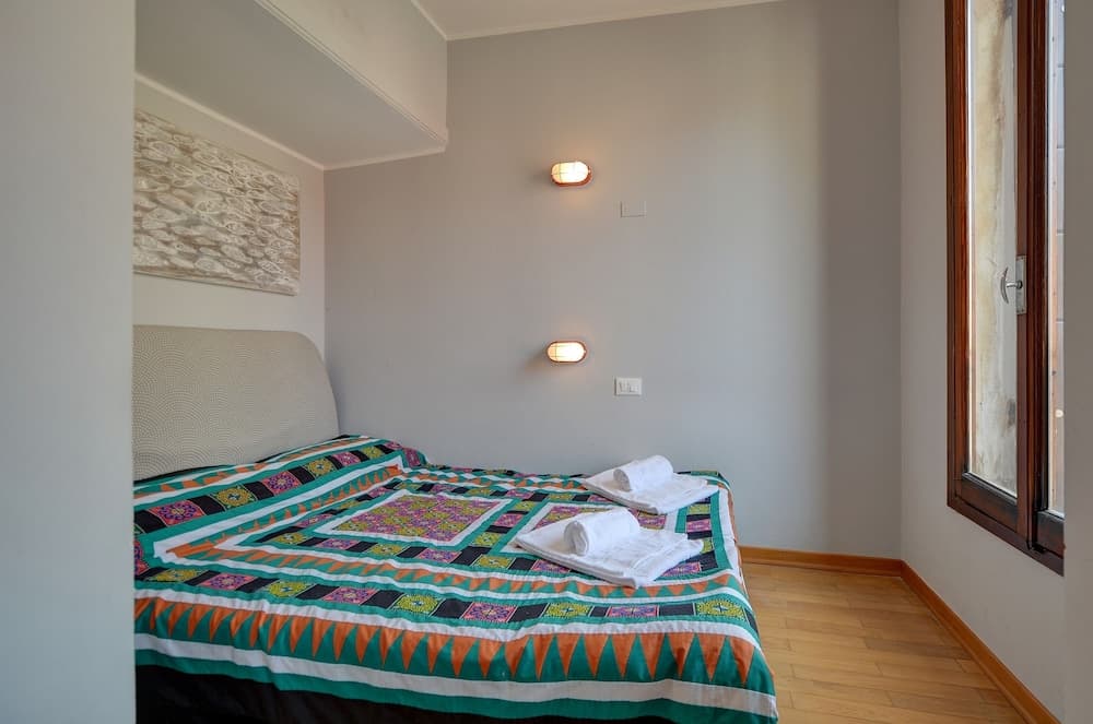 A cozy bedroom with a neatly made bed adorned in vibrant quilt patterns, soft lighting from wall-mounted lamps, wooden flooring, and a window letting in natural light, ideal for travelers seeking a comfortable and serene accommodation.