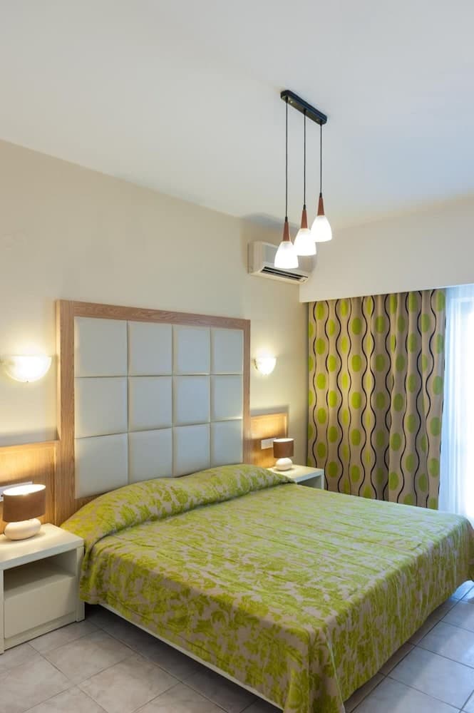 A cozy hotel bedroom features a neatly made bed with green patterned bedding, modern pendant lights, and stylish curtains, ideal for travelers seeking comfort and contemporary design.