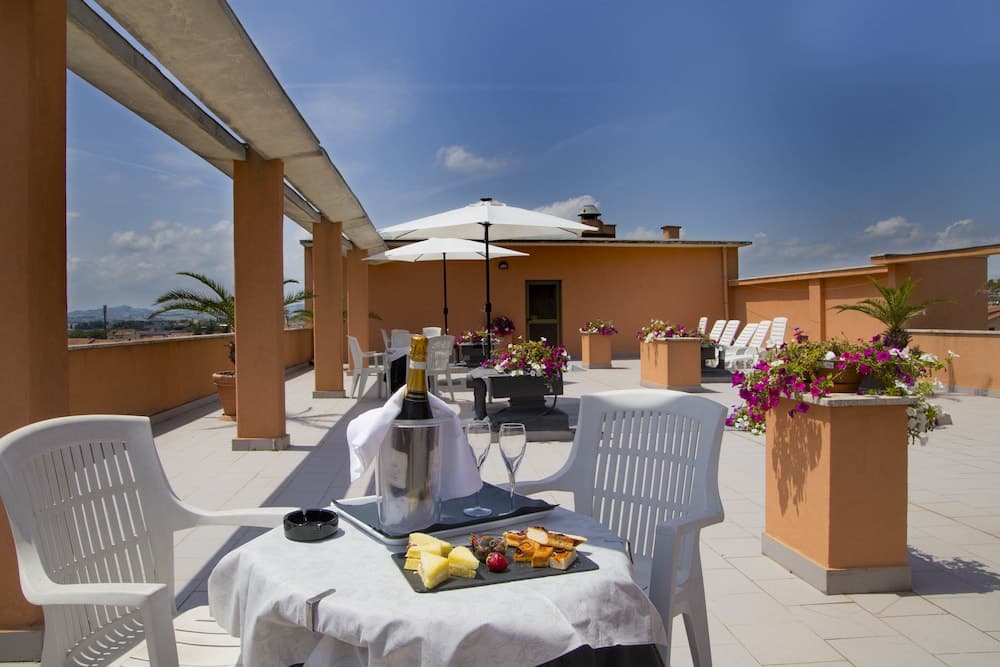 A serene rooftop terrace features white chairs, a table set with cheese and champagne, vibrant flower arrangements, and a panoramic view under a clear blue sky, ideal for a romantic getaway or leisurely relaxation.