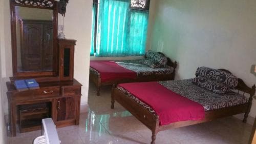 A cozy guesthouse room features two wooden beds with red bedding, a polished wooden dresser with a mirror, tiled floors, and green curtains, ideal for budget travelers seeking comfort.