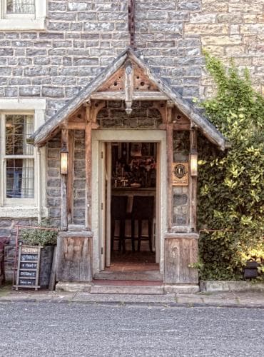 A charming stone cottage entrance with a rustic wooden porch invites travelers to explore its cozy interior, perfect for a quaint getaway in a serene village setting.
