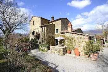 A serene Tuscan countryside villa with rustic stone architecture, lush greenery, terracotta pots, and a gravel courtyard invites travelers seeking a peaceful retreat in Italy's scenic landscapes.