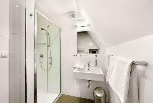 A modern, compact attic bathroom with white walls, a glass-enclosed shower, sleek sink, and mirrored storage exudes a clean and minimalist vibe, ideal for travelers seeking efficient space in boutique accommodations.