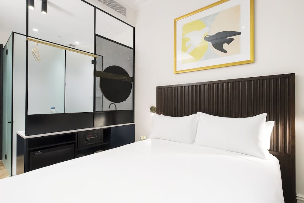 A modern hotel room features a cozy bed with white linens, a sleek wooden headboard, an abstract wall art piece, and a stylish mirrored wardrobe, ideal for travelers seeking comfort and contemporary design.