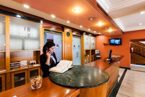 A warmly lit hotel reception area features a staff member on the phone behind a polished wooden desk, surrounded by shelves, a TV screen, and a cozy ambiance ideal for travelers seeking a welcoming check-in experience.