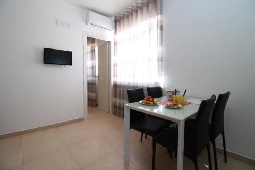 A cozy apartment dining area features a modern table set with colorful breakfast items, black chairs, a wall-mounted TV, and soft curtains, ideal for travelers seeking comfort and convenience in a compact living space.