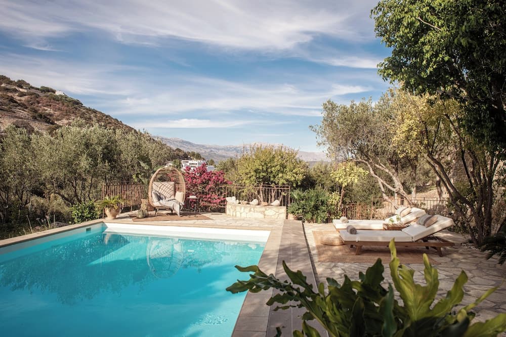 A serene outdoor poolside retreat surrounded by lush greenery and distant mountains, featuring cozy lounge chairs and a hanging egg chair, perfect for a tranquil vacation getaway.