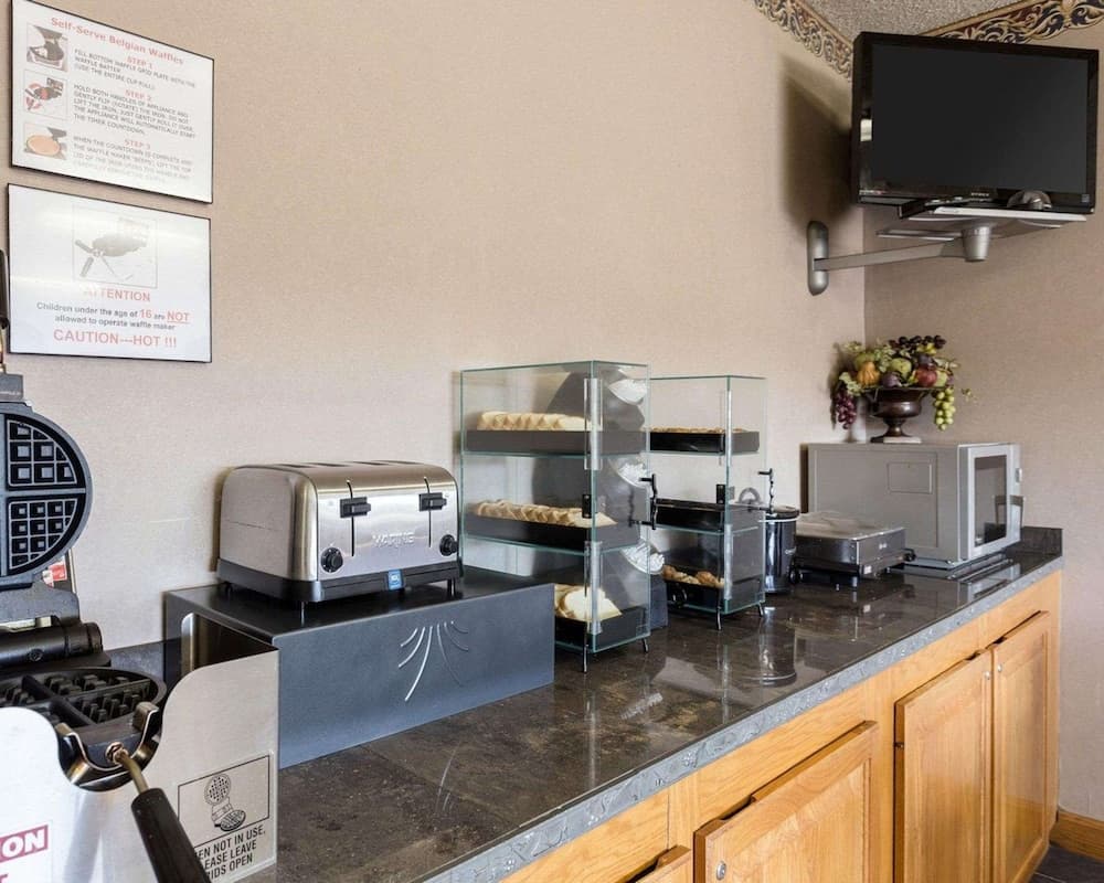 A cozy hotel breakfast area features a waffle maker, toaster, glass-covered pastries, and a microwave on a polished countertop, creating a welcoming atmosphere for travelers seeking a convenient morning meal.