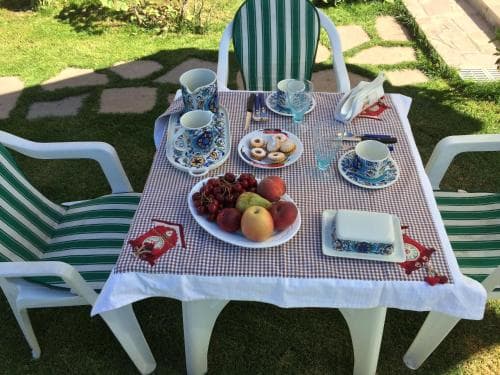 A serene outdoor breakfast setup on a grassy patio features a checkered tablecloth, colorful ceramic cups, fresh fruits, and pastries, inviting travelers to enjoy a peaceful morning in a cozy garden setting.