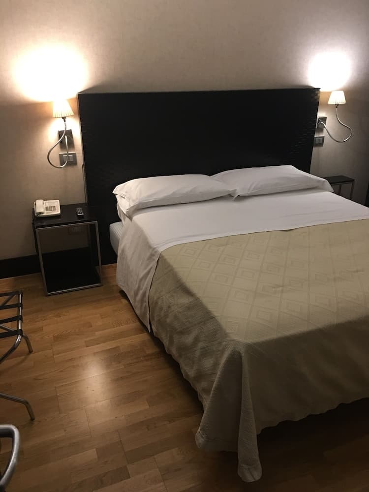 A cozy hotel room features a neatly made bed with white linens and beige accents, illuminated by warm wall-mounted lamps, ideal for travelers seeking comfort and relaxation.