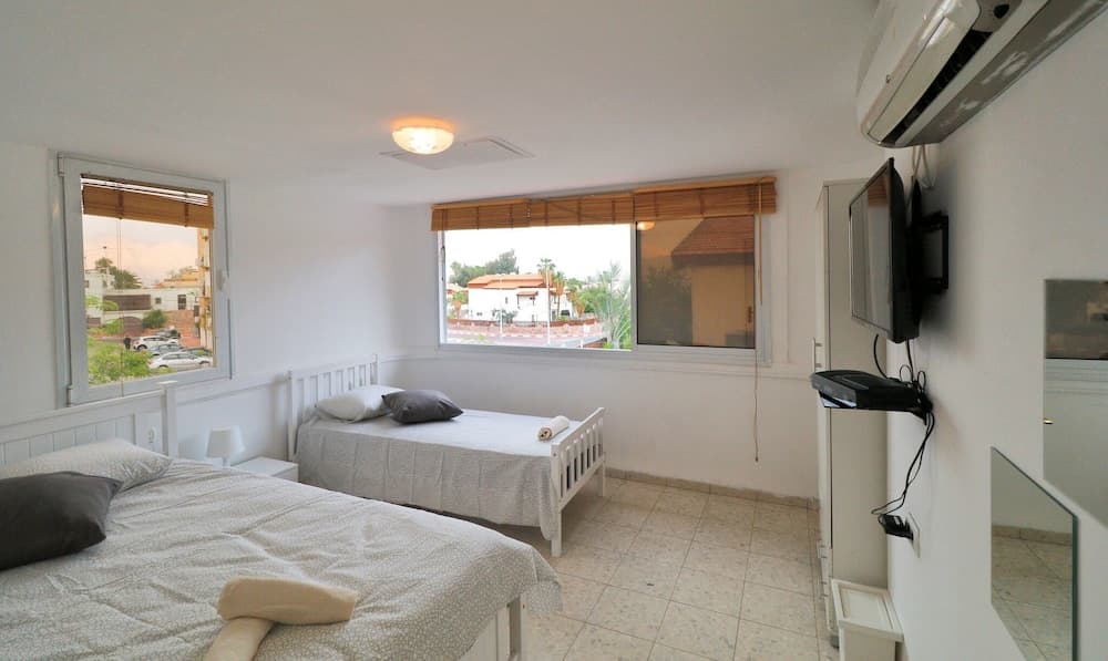 A cozy hotel room with two single beds, a wall-mounted TV, and large windows offering a serene view of palm trees and rooftops, ideal for travelers seeking comfort and relaxation.