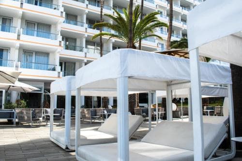 A serene hotel patio features white cabanas and lounge chairs under palm trees, ideal for relaxing travelers seeking a tranquil resort atmosphere.
