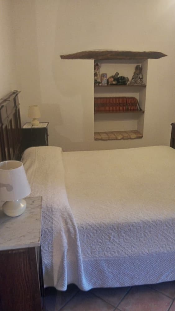 A cozy bedroom with a neatly made bed, wooden nightstands, white lamps, and a rustic alcove shelf displaying books and decor, ideal for travelers seeking a serene and charming accommodation experience.