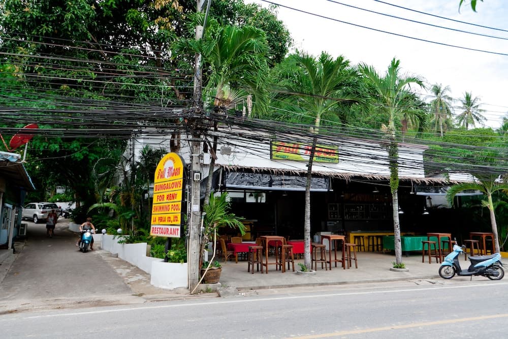 A vibrant tropical street scene featuring a restaurant with palm trees, outdoor seating, and a sign advertising bungalows for rent, ideal for travelers seeking a relaxed and casual dining experience in a lush environment.