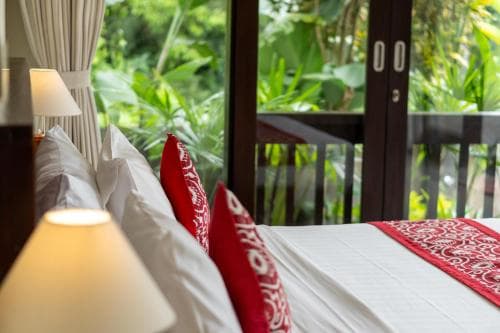 A cozy bedroom with white linens, red patterned pillows, and a warm lamp overlooks lush greenery through glass doors, ideal for travelers seeking relaxation in a tropical retreat.