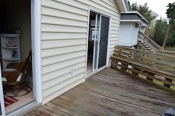 A cozy backyard deck with wooden flooring and railing invites relaxation, featuring a sliding glass door leading to a warmly lit interior with furniture, ideal for a serene getaway or home vacation setup.
