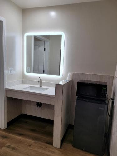 A cozy hotel room corner features a modern vanity with a bright LED-lit mirror, a sleek sink, and a compact fridge-microwave combo, ideal for travelers seeking comfort and convenience.