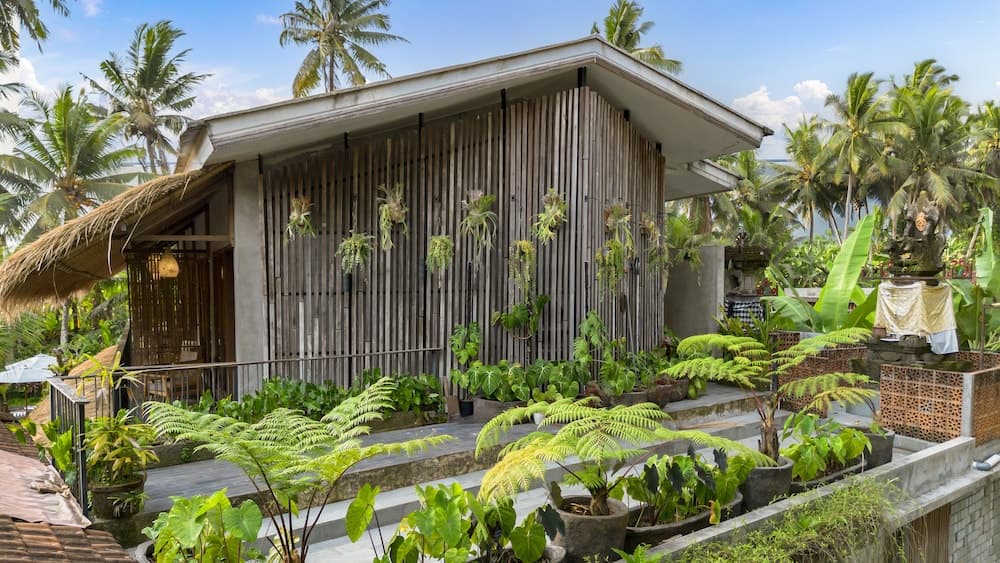 A serene tropical retreat features a wooden bungalow surrounded by lush greenery and palm trees, ideal for eco-conscious travelers seeking relaxation and connection with nature.