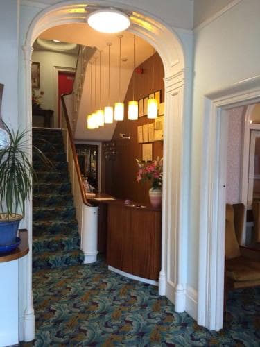 A warmly lit hotel lobby features an inviting staircase, wooden reception desk adorned with flowers, and cozy carpeting, ideal for travelers seeking a comfortable stay.