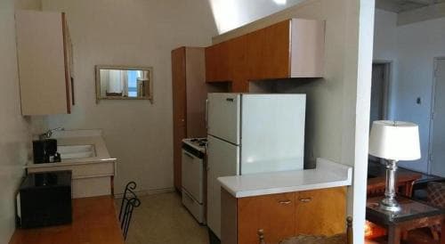 A cozy studio apartment features a compact kitchenette with wooden cabinets, a white refrigerator, and a sink, creating a warm and functional space ideal for short-term travelers seeking comfort and convenience.