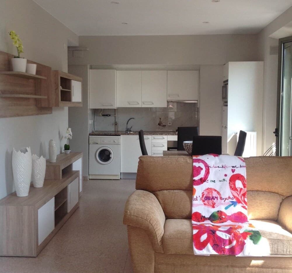 A cozy apartment living space features a beige sofa adorned with a colorful blanket, a modern kitchenette with white cabinets, and wooden shelves displaying decorative vases, ideal for travelers seeking a comfortable and stylish short-term rental.