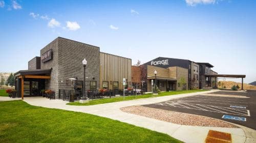 A modern commercial plaza featuring the "FORGE" building under a clear blue sky, surrounded by well-maintained greenery and outdoor seating areas, ideal for business travelers seeking a professional yet relaxed environment.