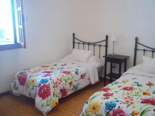 A cozy guesthouse room features two single beds adorned with vibrant floral bedding, a small nightstand with a lamp, and a window letting in natural light, ideal for travelers seeking a comfortable and inviting stay.