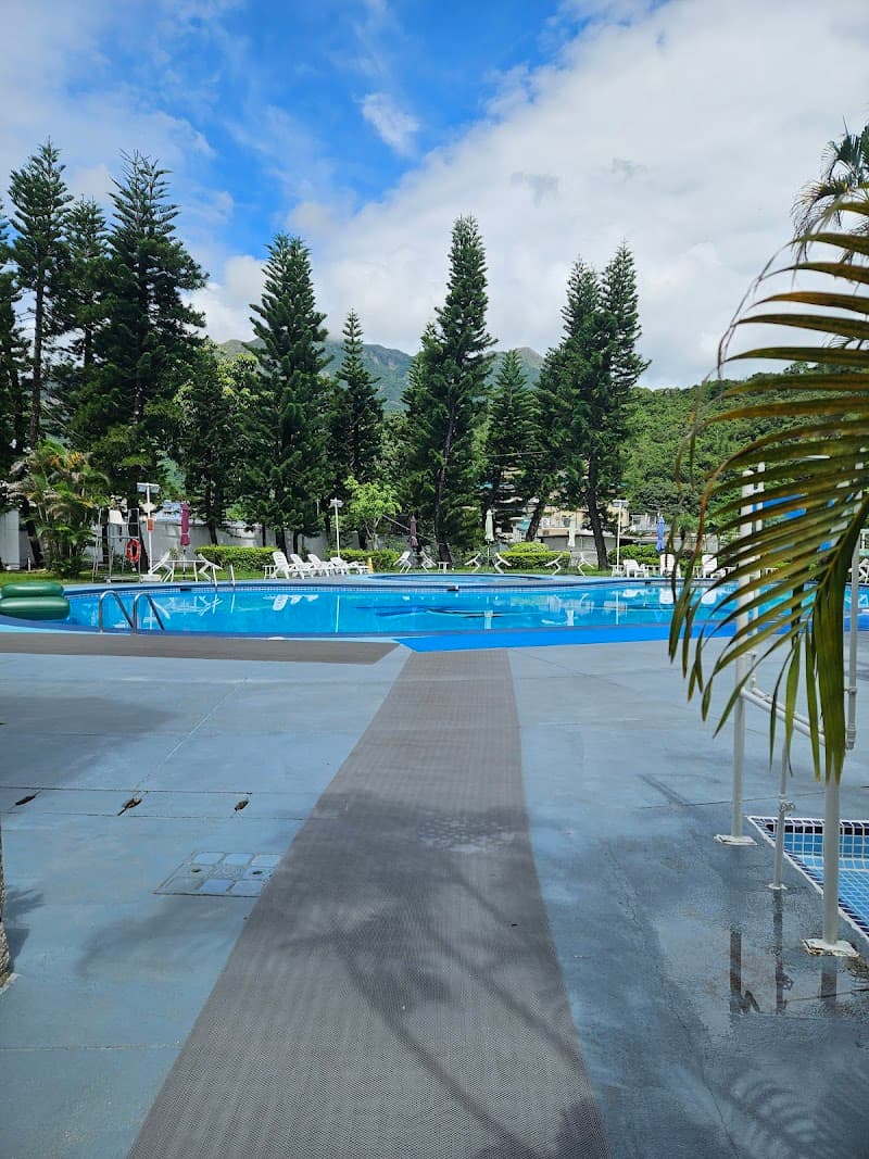A serene poolside scene at the Silvermine Beach Resort in Mui Wo, Lantau Island, Hong Kong, features a tranquil blue pool surrounded by lush green trees under a partly cloudy sky, ideal for relaxation and leisure travel.