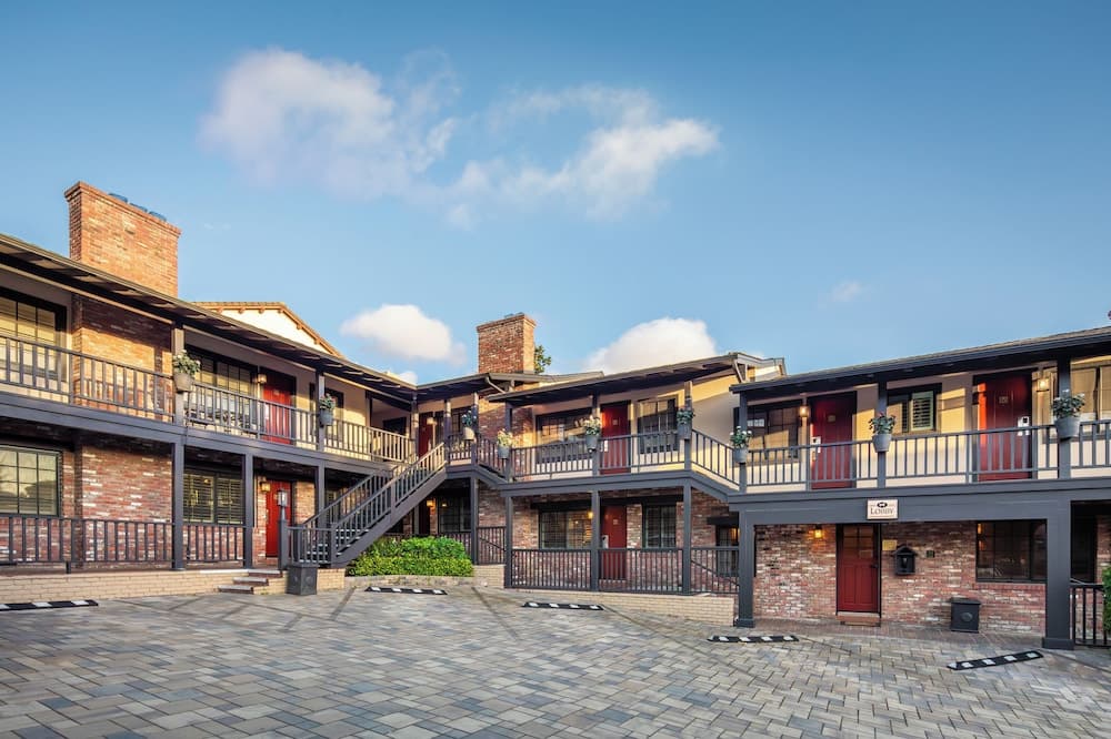 A charming brick courtyard hotel with red doors, balconies adorned with potted plants, and a clear blue sky creates an inviting atmosphere perfect for leisure travelers seeking a cozy stay.