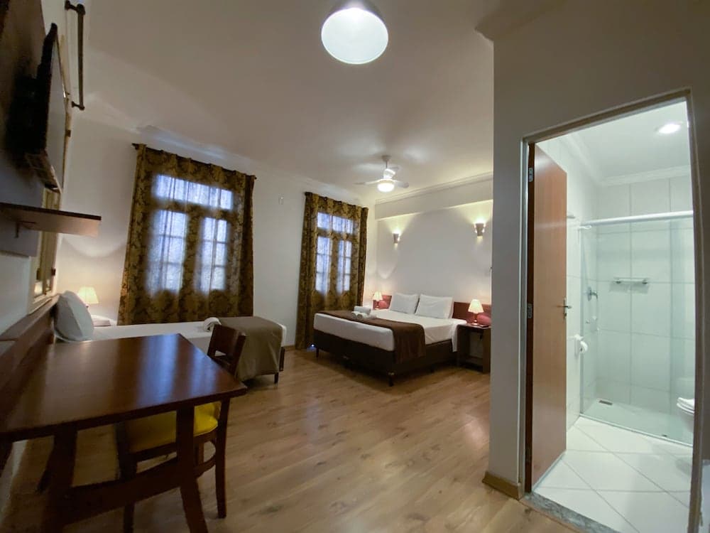 A cozy hotel suite features a warm wooden floor, elegant curtains, a comfortable bed, a dining area, and an adjacent bathroom with a glass shower, ideal for travelers seeking comfort and convenience.