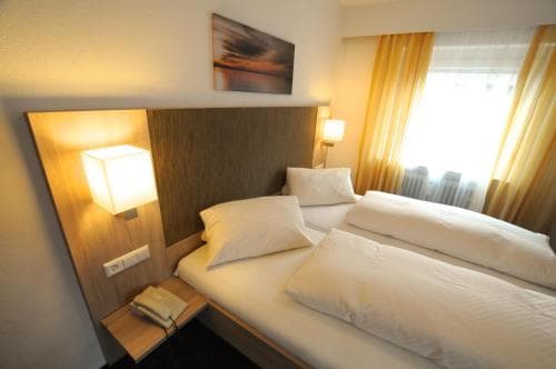 A cozy hotel room features a neatly made double bed with white linens, warm lighting from bedside lamps, and a serene view through sheer curtains, ideal for travelers seeking comfort and relaxation.