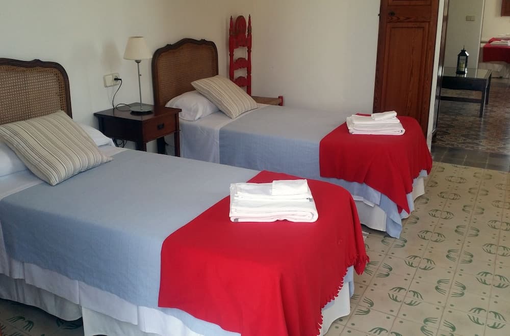 A cozy hotel room features two neatly made beds with blue linens and red accents, set against a patterned floor, ideal for travelers seeking comfort and simplicity.