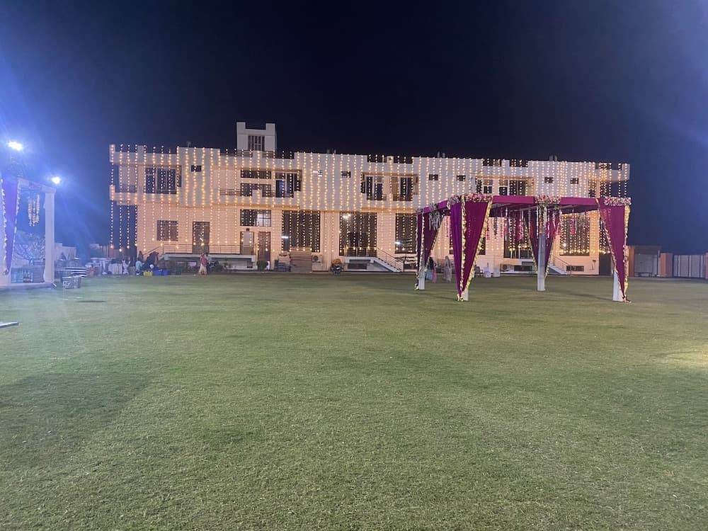 A festive outdoor event venue at night features a grand building adorned with twinkling lights, a lush green lawn, and elegant purple drapery structures, ideal for travelers seeking unique cultural celebrations or wedding experiences.