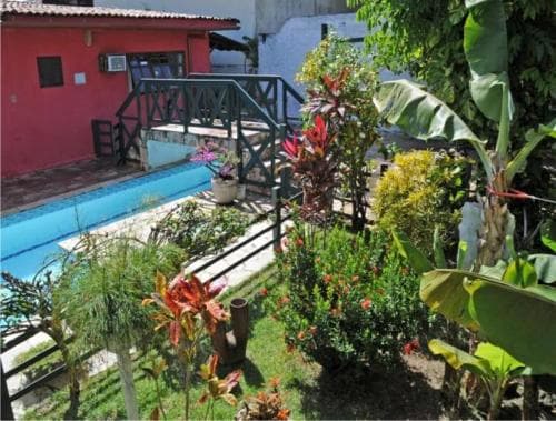 A serene tropical garden with lush foliage and vibrant flowers frames a tranquil poolside area beside a cozy red building, ideal for relaxing vacations in a private retreat setting.