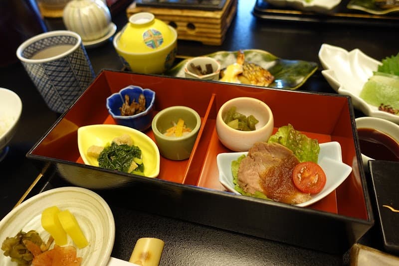 A beautifully arranged Japanese meal featuring colorful dishes in a red lacquer box, set on a dark table, evokes a serene dining experience ideal for travelers exploring culinary delights in Kyoto's Arashiyama district.