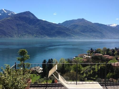 A serene lakeside view from a balcony showcases lush green hills, calm blue waters, and distant snow-capped mountains, ideal for tranquil retreats and scenic photography.