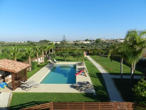 A serene tropical villa scene featuring a sparkling pool, lush greenery, palm trees, and lounge chairs under a clear blue sky, ideal for a relaxing vacation getaway.