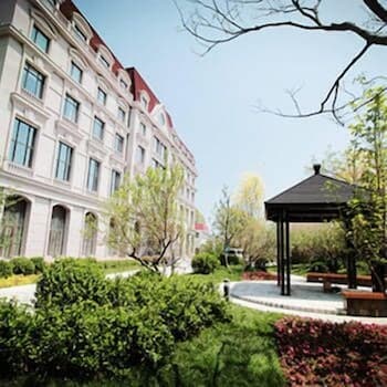 A serene urban garden with lush greenery, vibrant flowers, and a charming gazebo invites relaxation near a grand multi-story building, ideal for travelers seeking a peaceful retreat in a city setting.