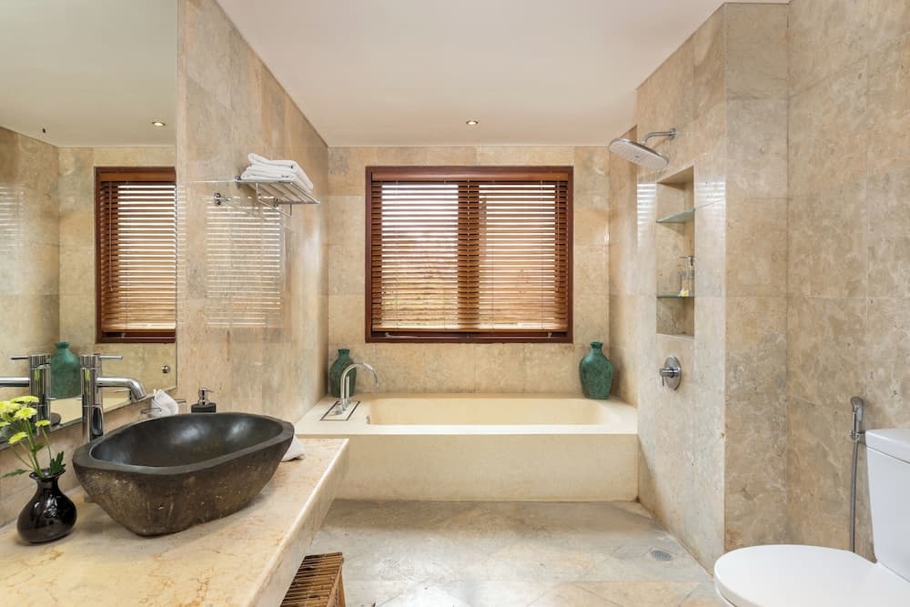 A serene luxury bathroom with beige marble walls, a sunken bathtub, a stone sink, wooden blinds, and decorative vases, ideal for a relaxing retreat during a high-end resort stay.
