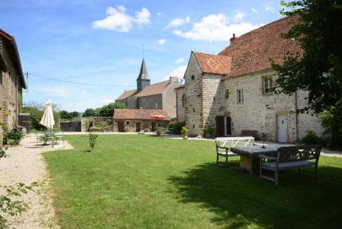 A serene rural courtyard features stone buildings, a grassy lawn, outdoor seating, and a distant church spire under a partly cloudy sky, ideal for a peaceful countryside retreat.