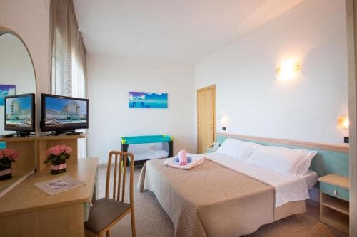 A cozy hotel room features a neatly made bed, a desk with two monitors, and a vibrant painting on the wall, creating a welcoming atmosphere ideal for travelers seeking comfort and convenience.