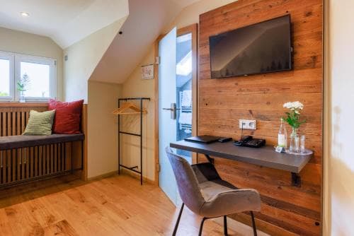 A cozy attic-style hotel room with wooden walls, a desk setup featuring a TV and flowers, and a bench by the window, ideal for travelers seeking comfort and functionality in a modern retreat.