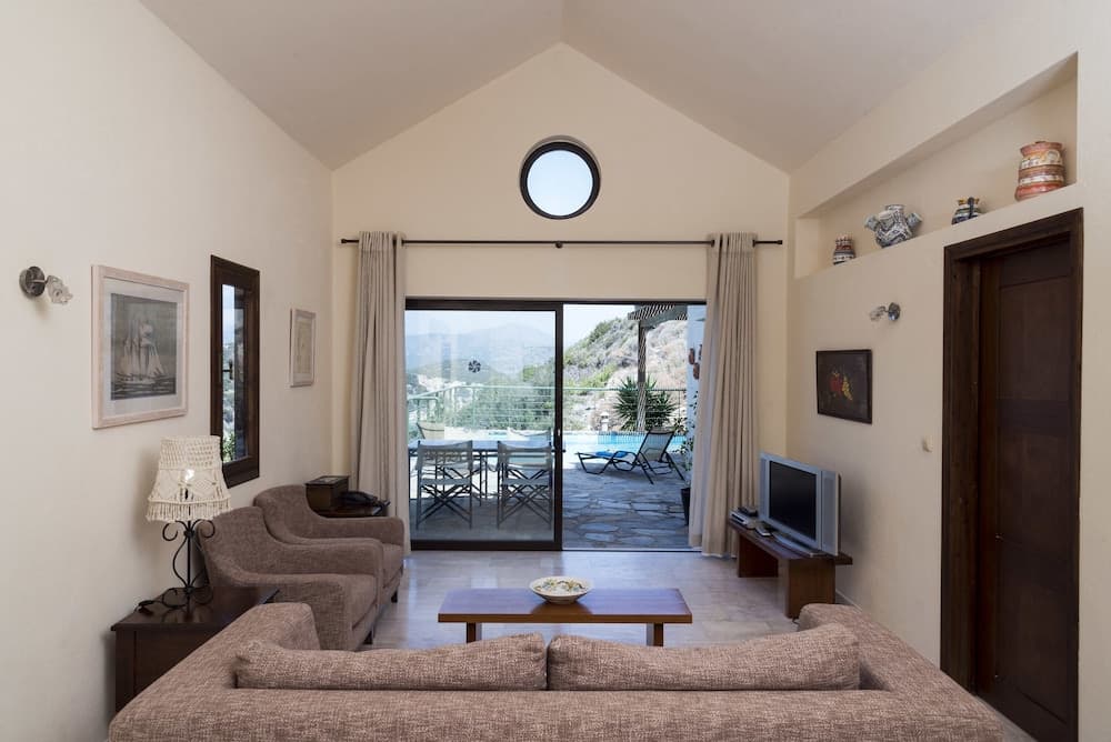 A cozy living room with beige walls, a vaulted ceiling, and a round window opens to a serene outdoor terrace with lounge chairs and a pool, ideal for a relaxing vacation retreat.