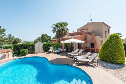 A serene Mediterranean villa features a sparkling pool, sun loungers under an umbrella, lush greenery, and a clear blue sky, ideal for a relaxing family vacation.