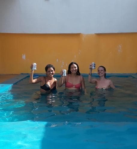 Three women enjoying drinks in a vibrant blue pool against a yellow wall create a joyful and relaxed atmosphere, perfect for a fun-filled vacation photo.